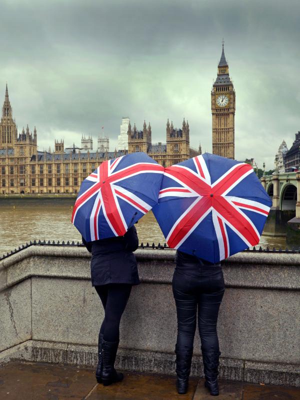 Two ladies standing with Union Jack umbrellas in front of the Houses of Parliament in London.