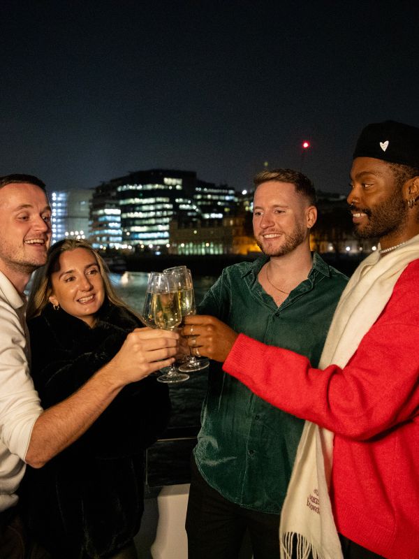 Four friends raising glasses on the deck of a Thames evening cruise, with the City of London skyline behind them.