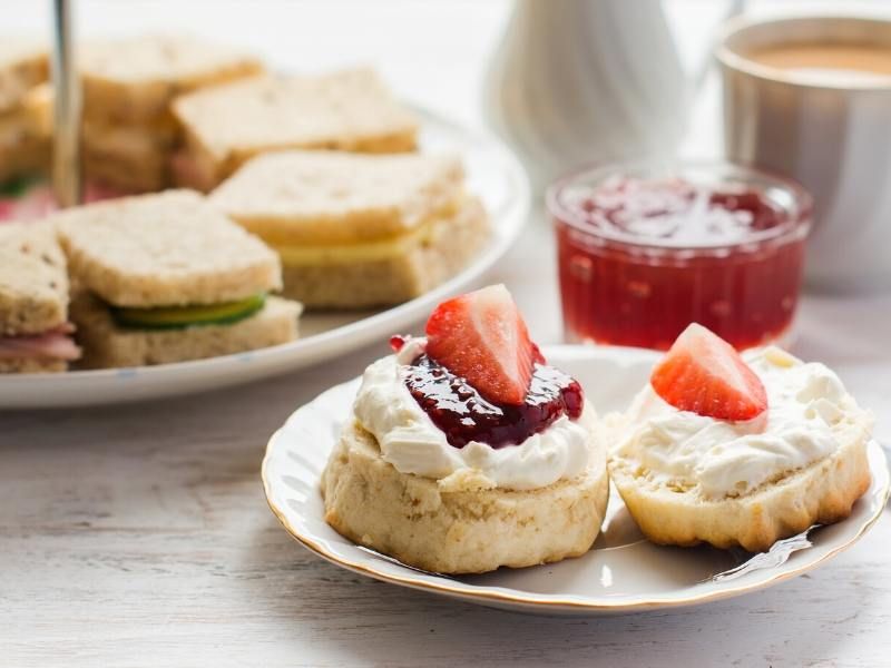 A plate with a scone, jam and cream with a plate of sandwiches in the background