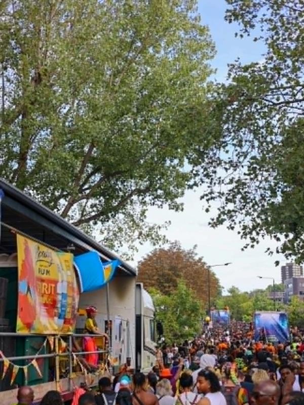 Crowds filling a tree-lined street at Notting Hill Carnival in London, with a sound system truck and rum banner overhead