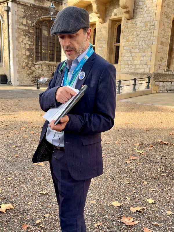 Blue Badge guide in a flat cap with an Institute of Tourist Guiding lanyard, speaking to the group in the Tower of London
